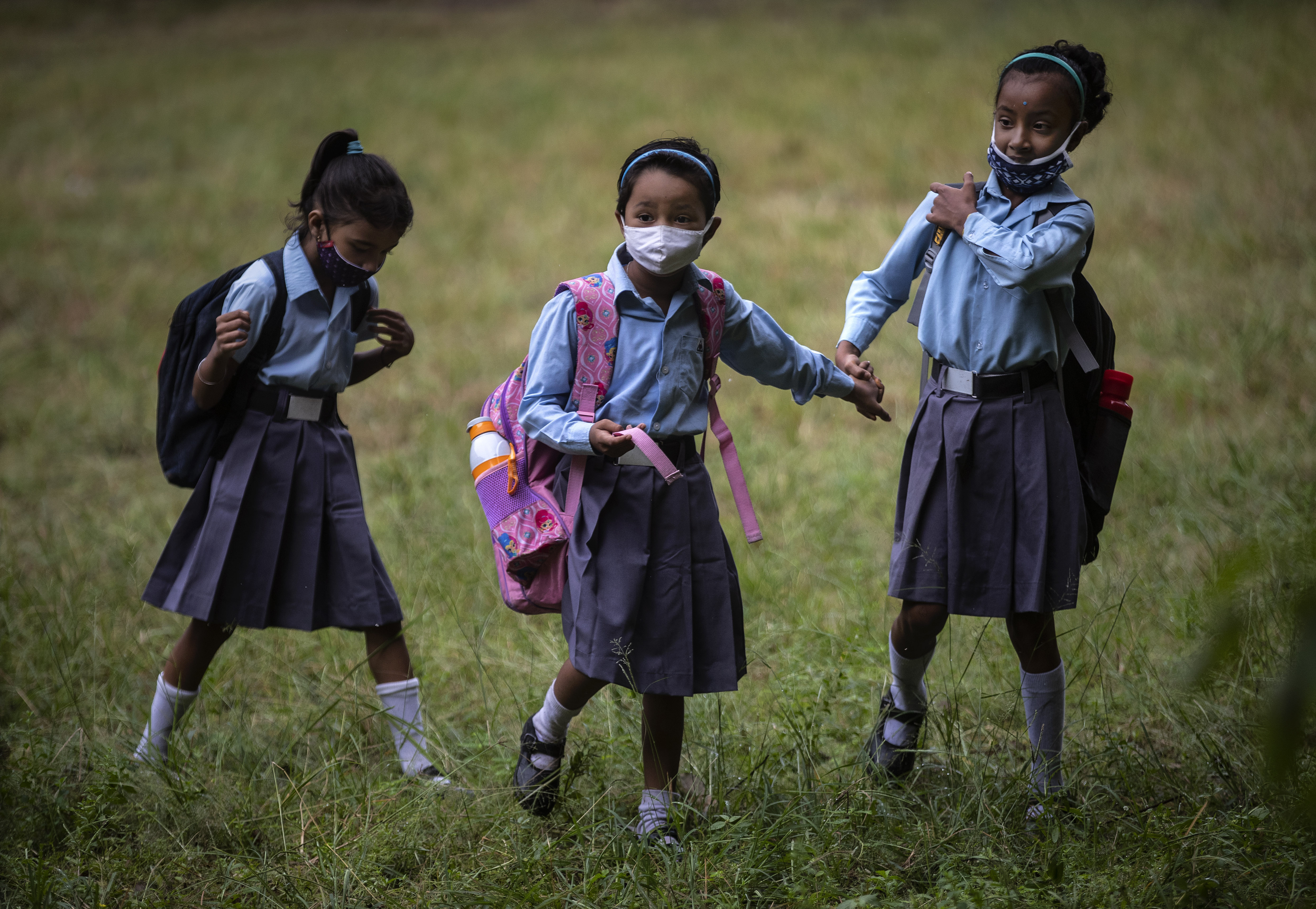 Three young girls stand in a field, wearing navy blue uniforms and face masks. 